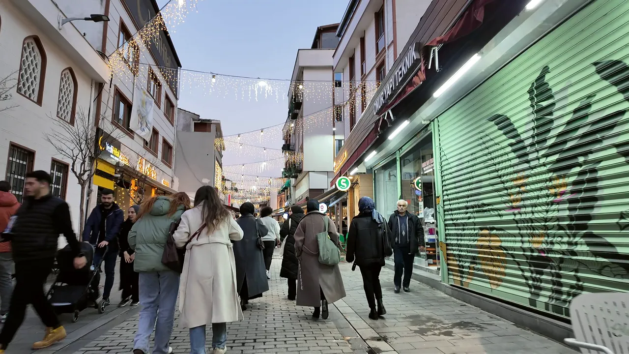 A street at dusk under festoon lights, shopfronts open and people walking.